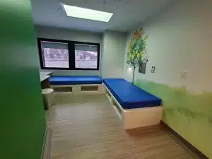 Inside of a room at the mental health unit at Children's Minnesota that has two beds, a desk, and a large window facing trees. 